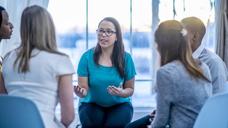 A multi-ethnic group of adults are attending a mental health support group. A woman is sharing her problems while they sit in a circle.